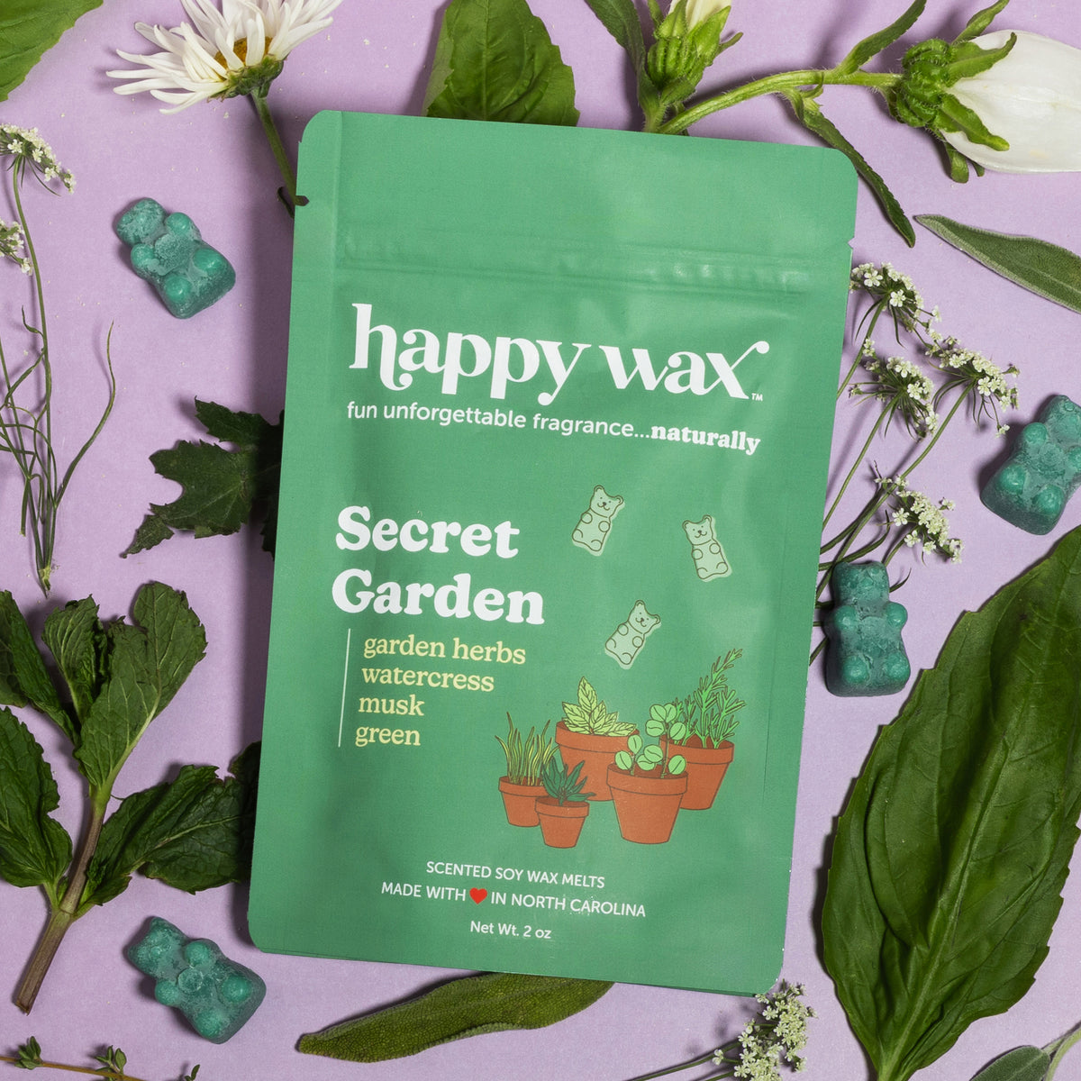 Green pouch of Happy Wax Secret Garden soy wax melts surrounded by fresh herbs and flowers on purple background.