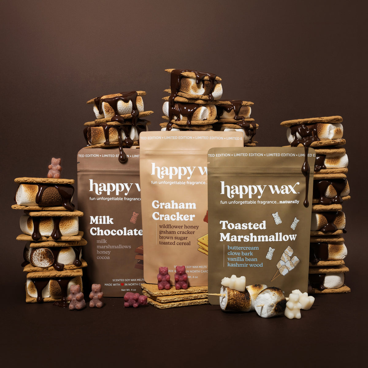 Three Happy Wax candle boxes labeled Milk Chocolate, Graham Cracker, and Toasted Marshmallow, surrounded by stacked s'mores-t