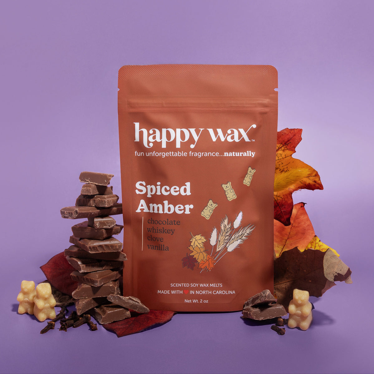 Brown pouch of Happy Wax Spiced Amber wax melts surrounded by stacked chocolate pieces, autumn leaves, and cream-colored wax 