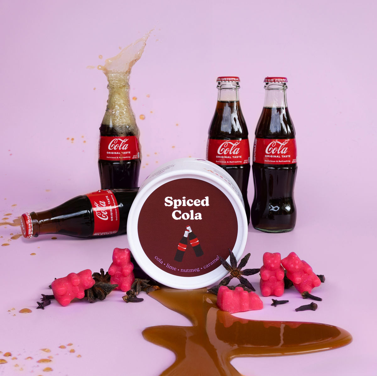 White container of Spiced Cola wax melts surrounded by cola bottles, pink berries, and spice garnishes on pink background.