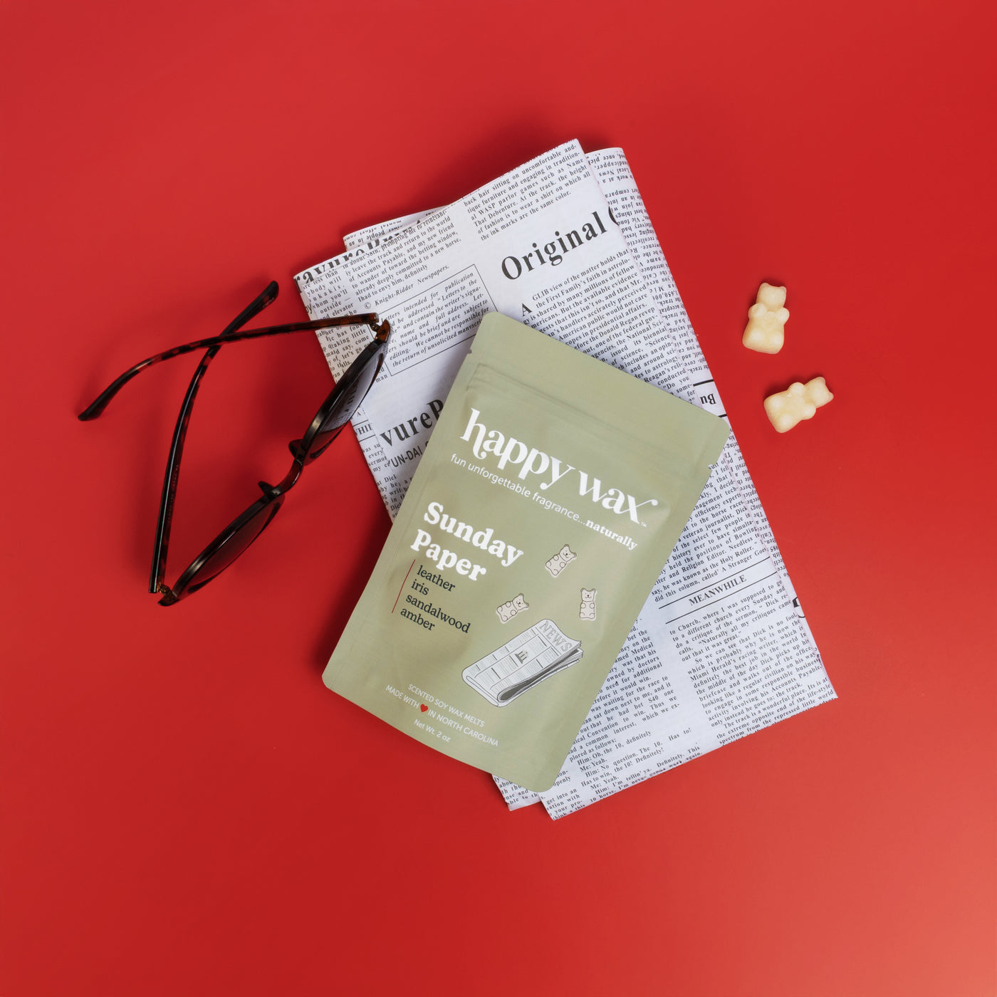 Pale green Happy Wax Sunday Paper pouch on newspaper clippings with black glasses and cream-colored wax melts on red backgrou