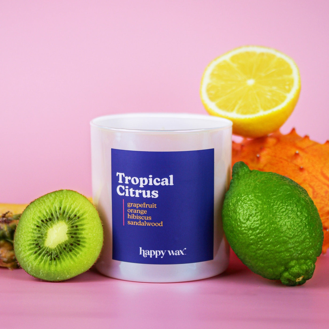 White candle with navy label surrounded by lemon slice, kiwi, lime, and orange against pink background.