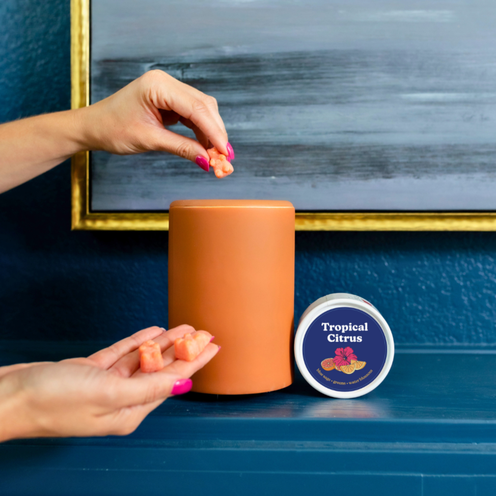 Hand holding orange wax melts above cylindrical container next to Tropical Citrus labeled tin.