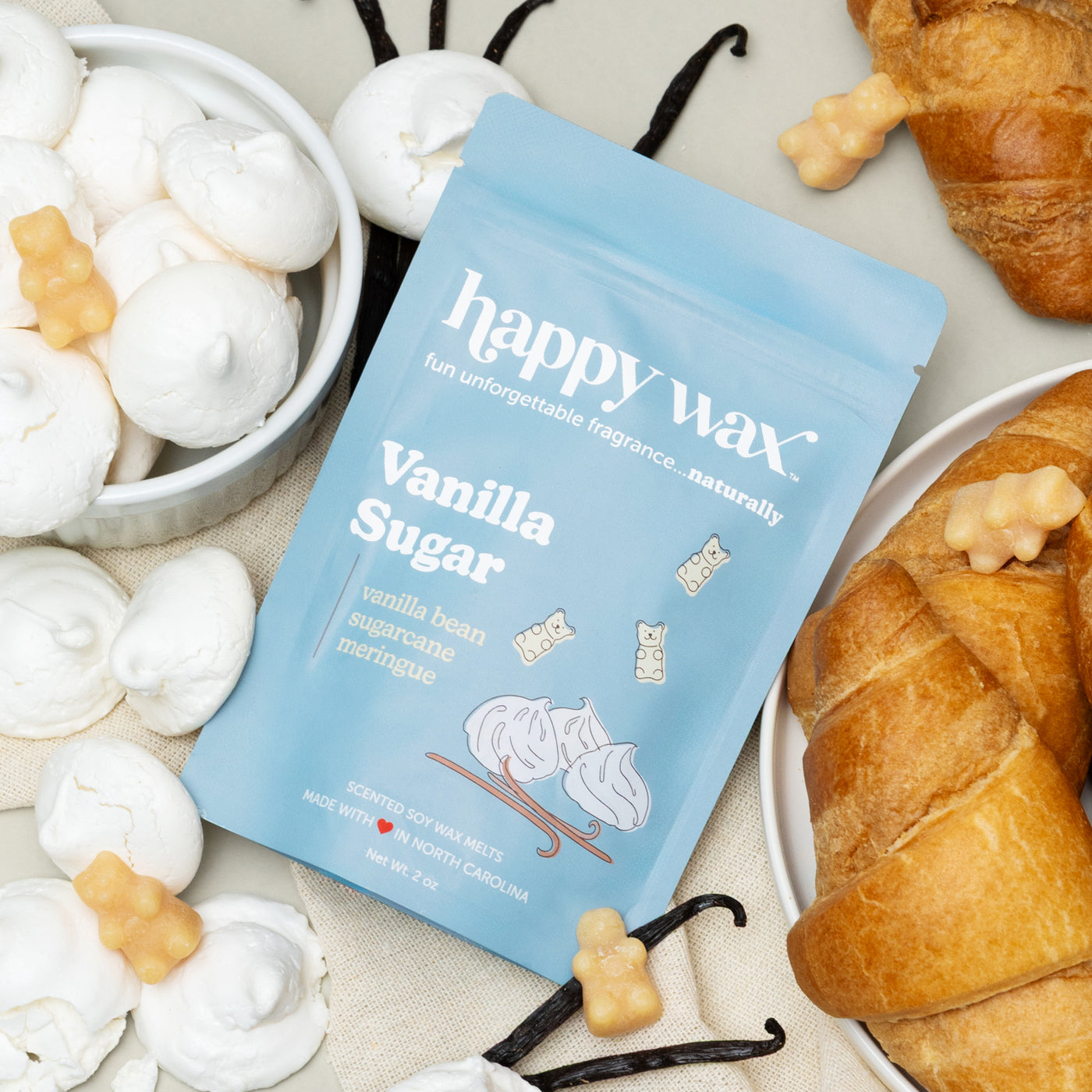 Blue "happy wax" Vanilla Sugar wax melt package surrounded by white wax melts, vanilla beans, pastries, and bear-shaped wax p
