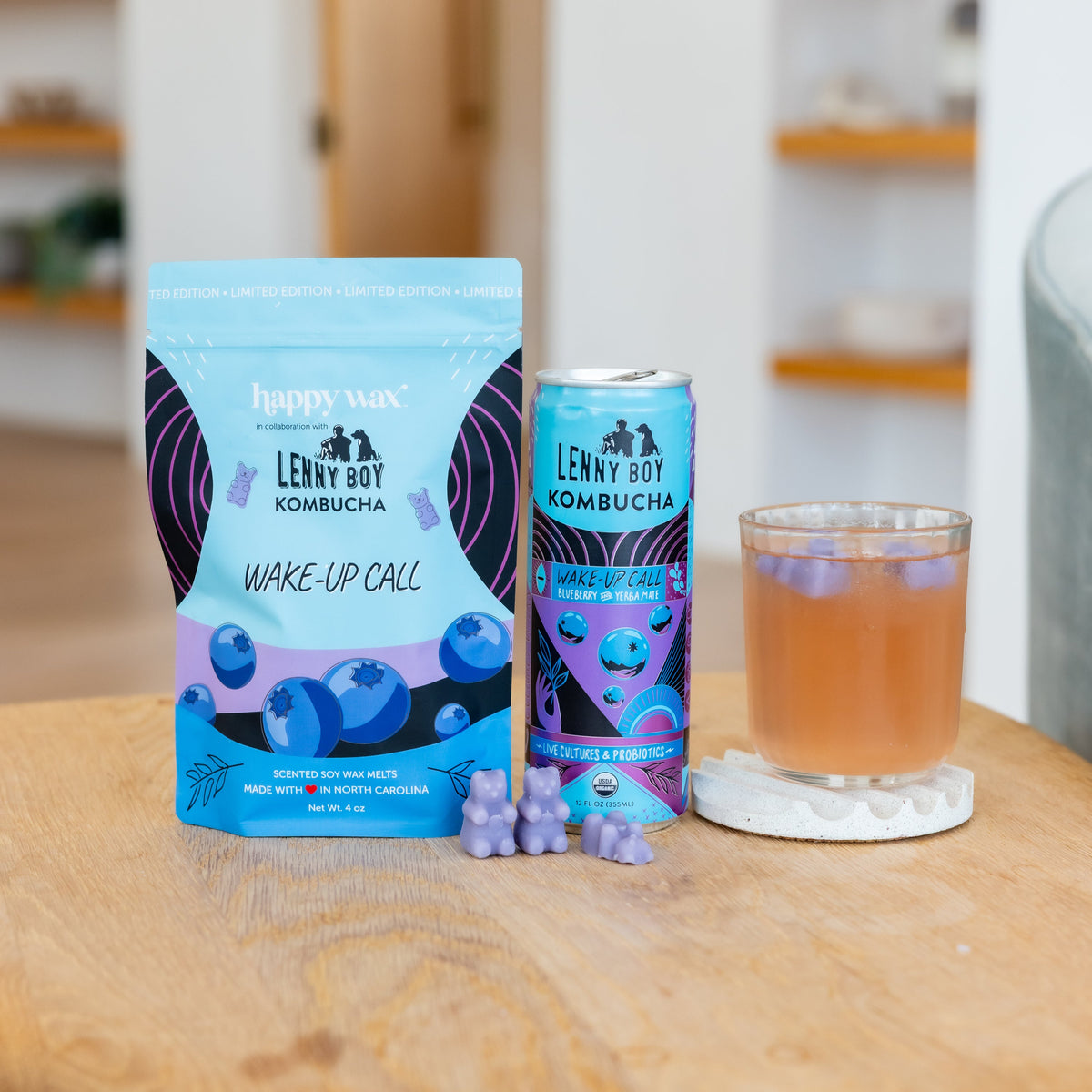 Blue and purple wax melts package, kombucha beverage can, glass with amber liquid, and purple wax melts on wooden surface.