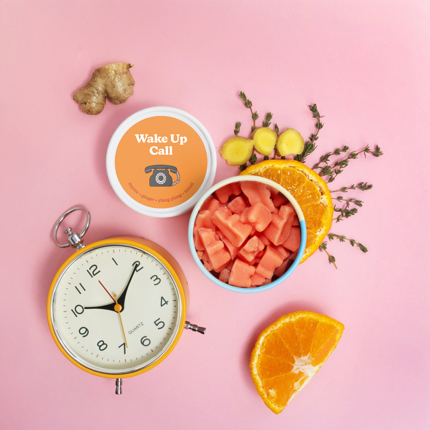 Two white bowls of wax melts surrounded by an alarm clock, ginger root, citrus slices, and dried herbs on pink background.