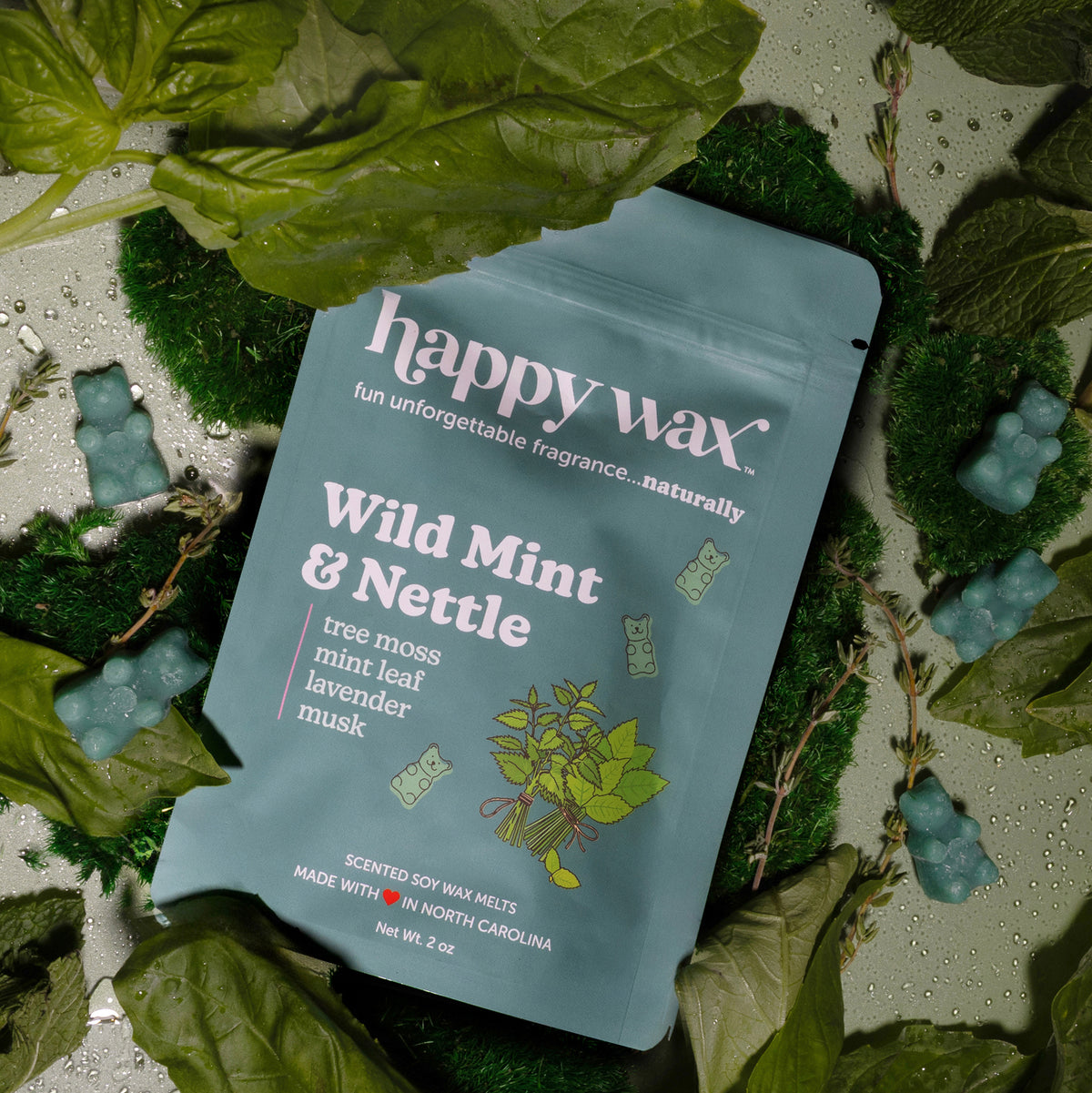 Gray pouch of Happy Wax Wild Mint & Nettle wax melts surrounded by fresh mint leaves, moss, and scattered wax melts on white 