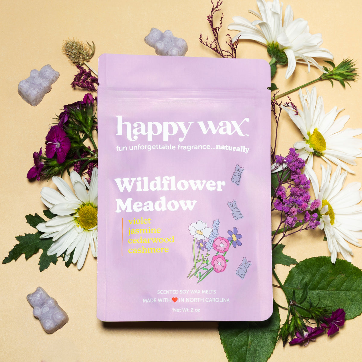 Lavender pouch of wax melts surrounded by purple cloud-shaped melts, white daisies, purple flowers, and dried botanicals on b