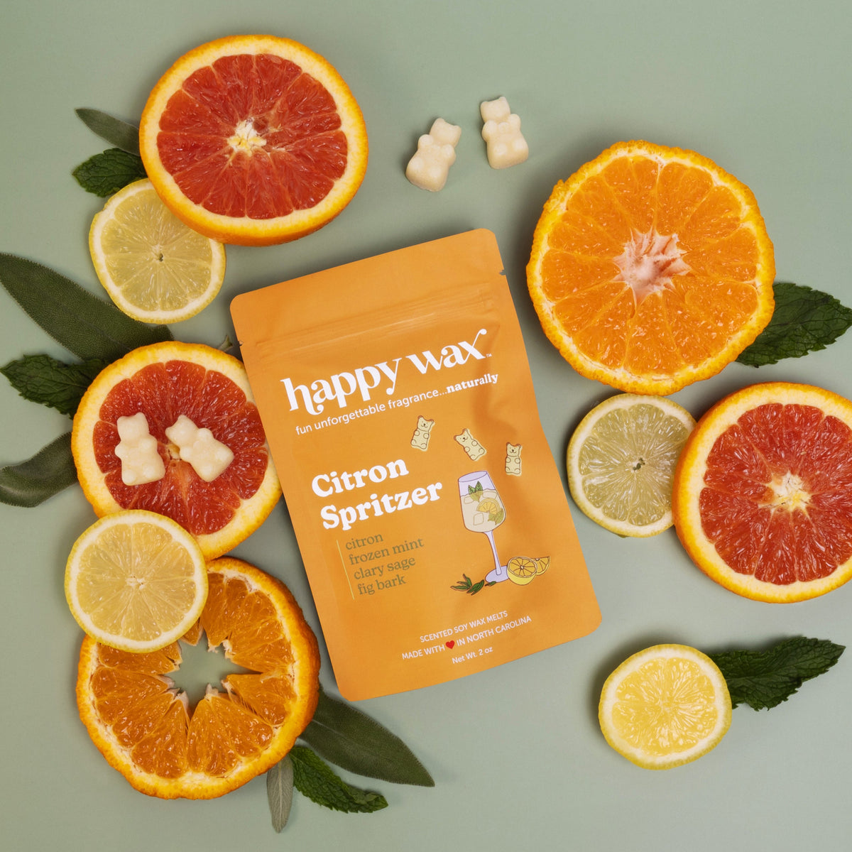 Orange pouch of Happy Wax Citron Spritzer wax melts surrounded by fresh citrus slices and two cream-colored wax melts.